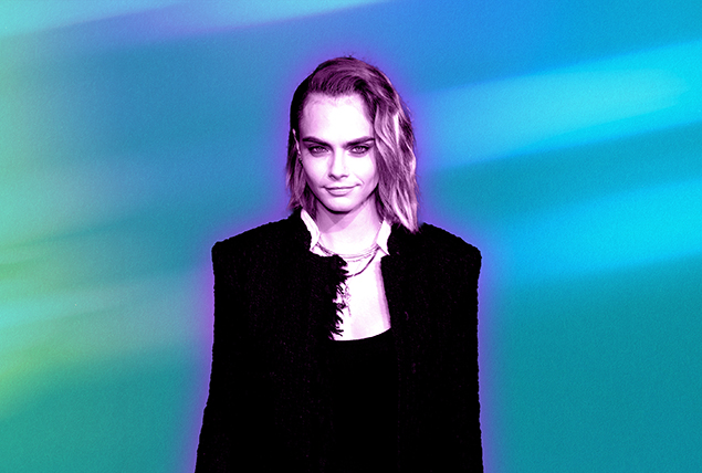 Cara Delevingne stands in a black dress against a teal background.