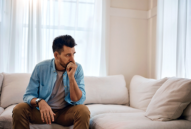 A man sits on a sofa with his hand under his chin looking to the side in throught.