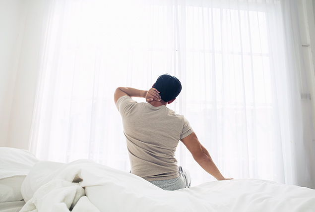 A man sits in bed in the morning reaching up to his neck and stretching.