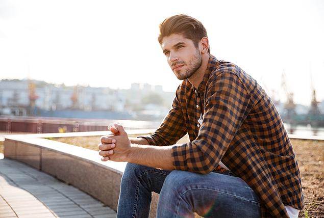 A man in a checkered shirt sits outside on a small wall with his hands clasped together.