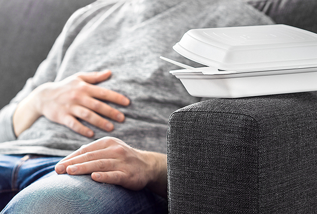 A man sits on a couch holding his stomach with a takeout container sitting next to him.