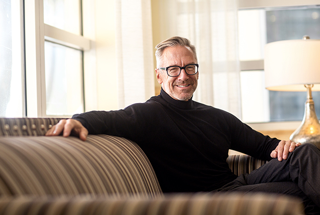 A man over 65 years old sits on a sofa smiling.