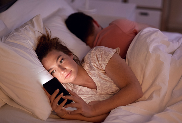 A woman lays in bed looking at social media while her male partner sleeps.