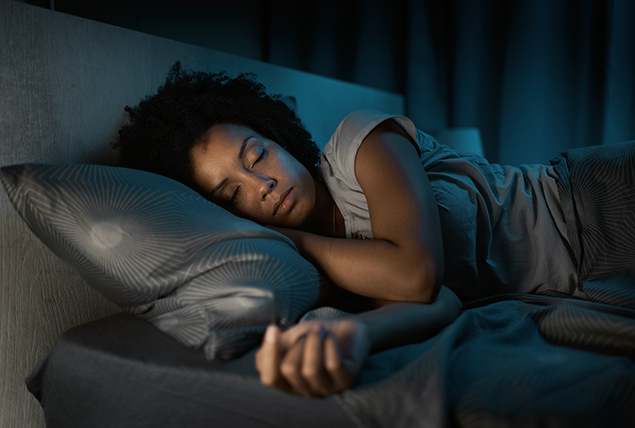 A woman lays asleep on her bed next to a dimly lit lamp on her bedside table.