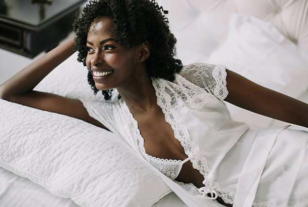 A black woman lays down on the bed smiling with her head propped up on her arm.
