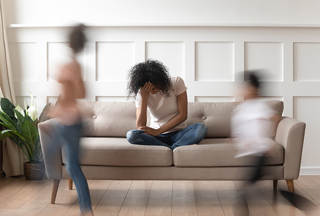 A woman sits on a chair with her head in her hand while two kids run around the room.