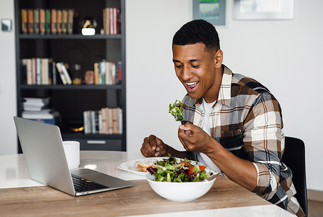 A man sits at his laptop eating a salad.