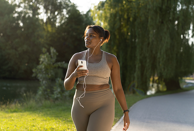 A black woman in activewear walks along a path outside while listening to music.