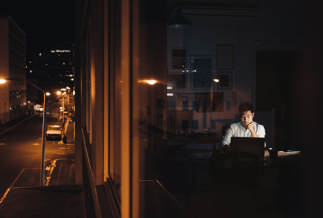 A man is seen through a city window working at his laptop late at night.
