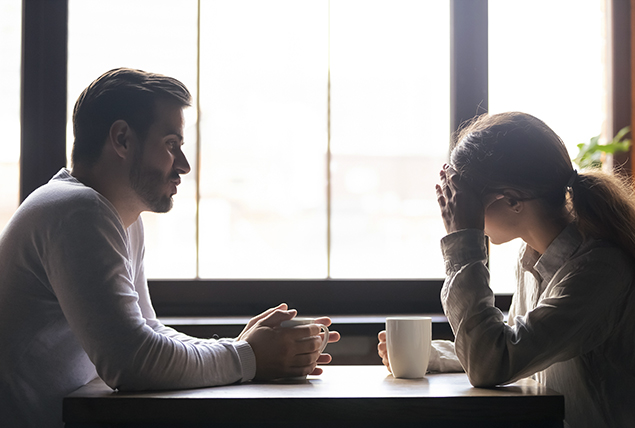 A man sits across from a woman who is holding her head with one hand and a cup of coffee in another.