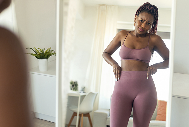 A woman in activewear stands in front of a mirror looking at her body.