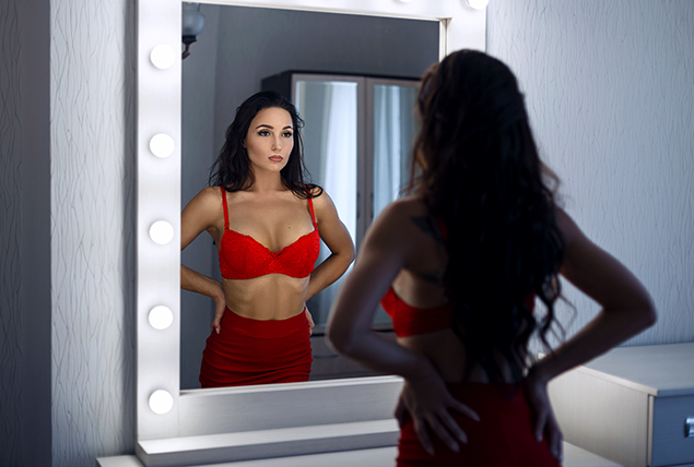 A woman stands in front of a mirror in a skirt and bra top with her stomach sucking in.