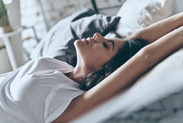 A woman lays down with her eyes closed as she reaches over her head.