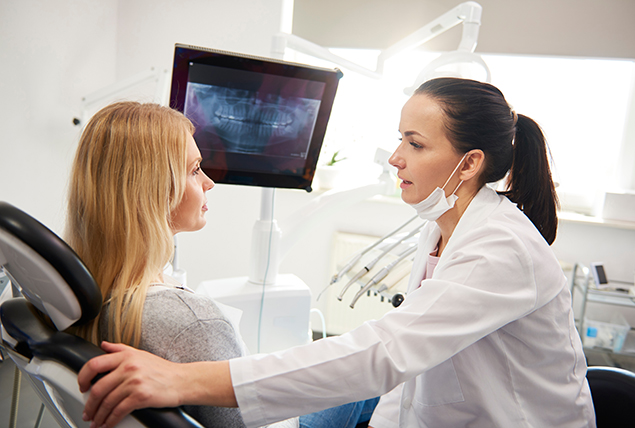 a woman in a dentist office talks with a dentist while an x ray is displayed on a screen