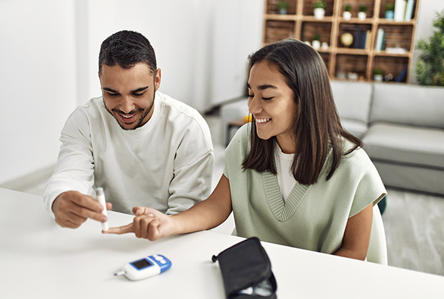 a man and woman sit at kitchen counter and check her blood sugar on her fingertip