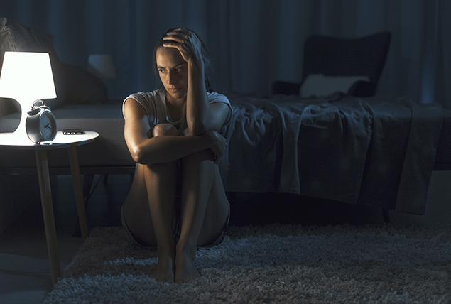 stressed woman leans on side of the bed while looking at alarm clock