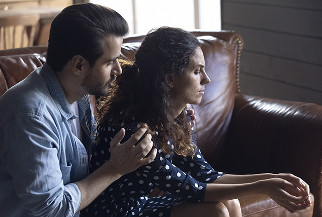 a man holds a stressed woman as they sit on a brown leather couch