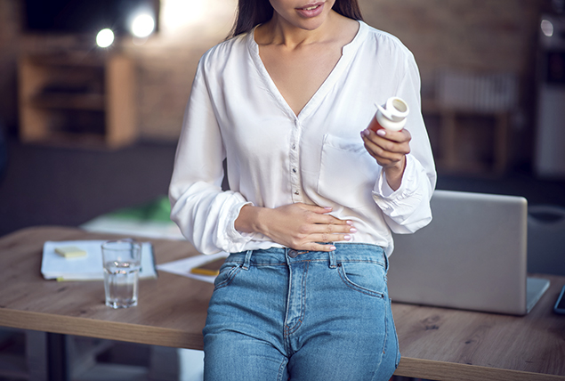 woman in white shirt holds stomach while looking at pill bottle