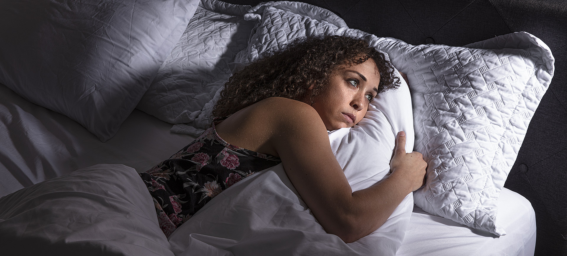 woman lays in bed with white sheets and hugs white pillow looking stressed