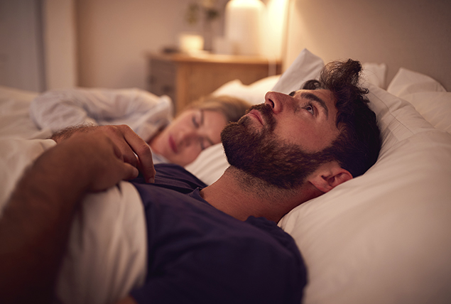 man lays in bed and stares at ceiling as woman sleeps next to him 