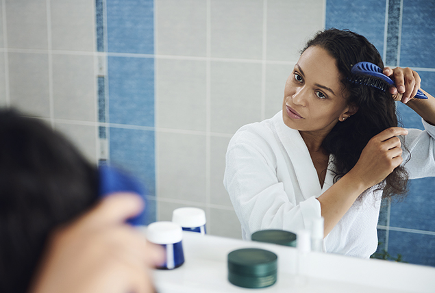 woman of color in white bathrobe stands in mirror and brushes her hair