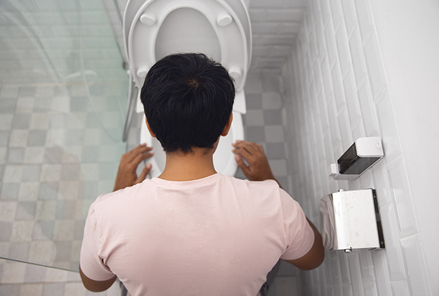 man in light pink shirt leans over toilet bowl in a gray tile bathroom
