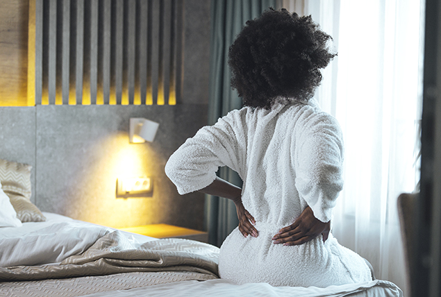 woman in white bathrobe sits on edge of bed and holds her lower back in pain