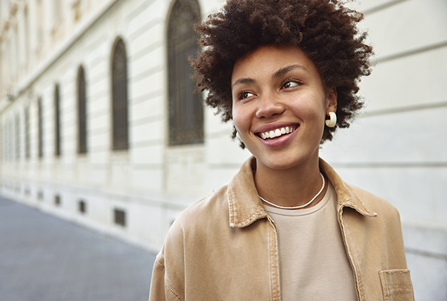 woman in beige shirt walks along white brick wall and smiles