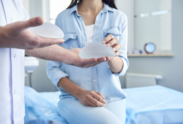 doctor holds two breast implants as woman seats on doctors table and reaches for one