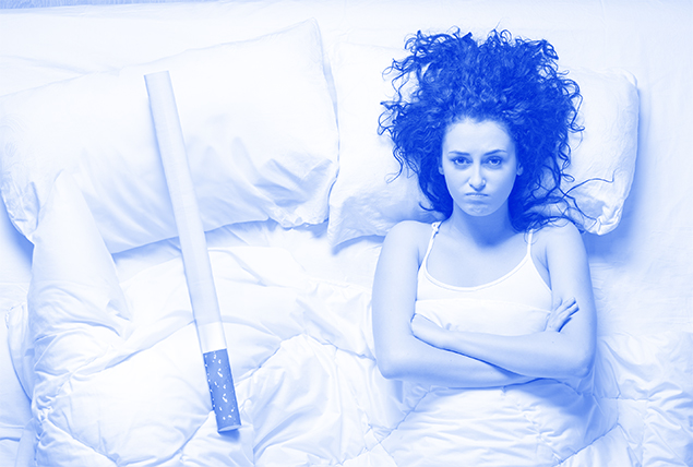 woman lays in bed next to giant cigarette with her arms crossed over her chest looking angry with blue tint
