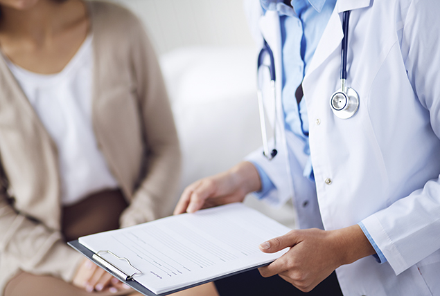 doctor in white coat holds clipboard and talks with female patient in tan cardigan