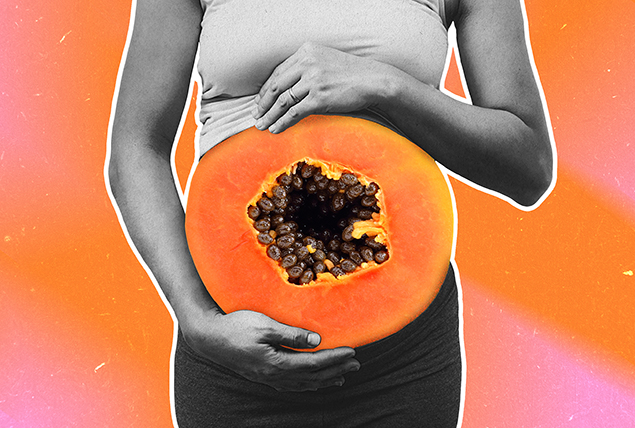A woman holds her pregnant stomach with half of an open-faced papaya over her belly.