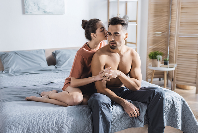 man and woman sit on bed holding hands as she is about to kiss his neck and he looks disinterested