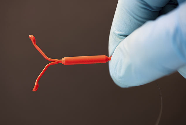 blue gloved hands hold red IUD on brown background