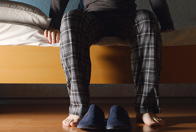 man's legs in plaid pj pants sitting on side of bed with slipper on the floor in front