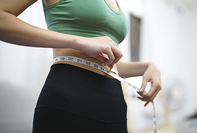 A woman uses measuring tape around her waist while dressed in workout gear.