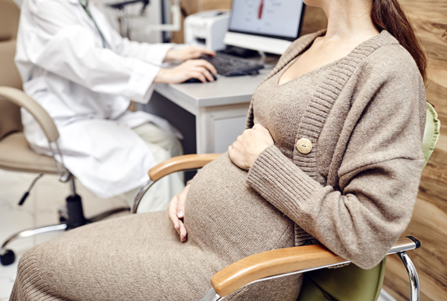 pregnant woman in beige dress holds her belly sitting in doctor's office with doctor in white coat sitting nearby