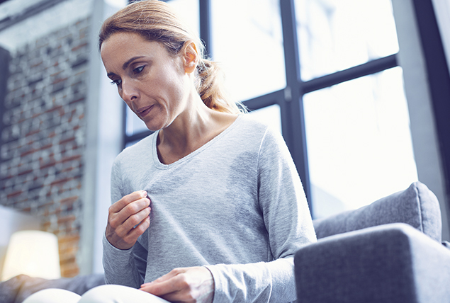 woman in long sleeved gray top tugs at her top trying to cool herself down