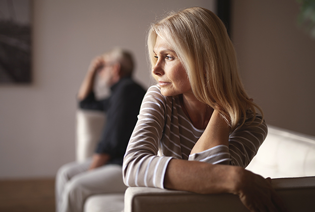 woman in striped shirt looks stressed and sits in couch as man sits in the distance