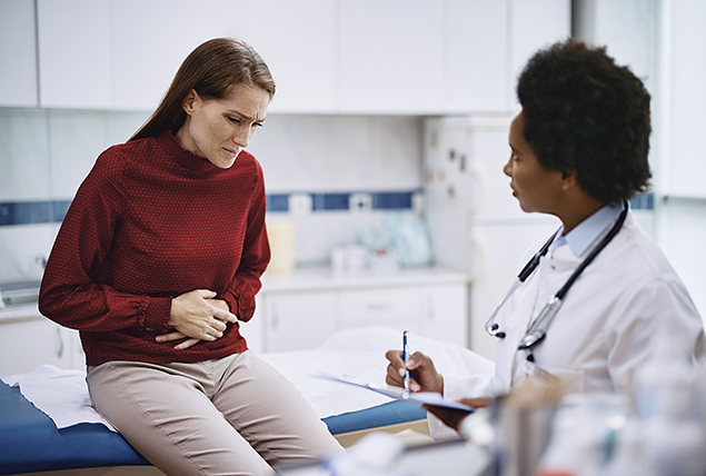 A woman in a red sweater holds her abdomen as she sits on a bed talking to a doctor.