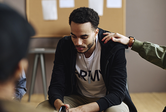 man in army shirt hangs his head sitting in support circle group as someone touches his shoulder