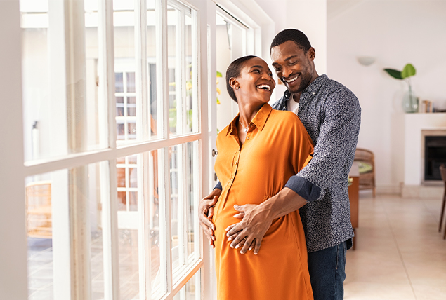 pregnant woman in orange dress smiles at man as he holds her from behind also smiling touching her belly