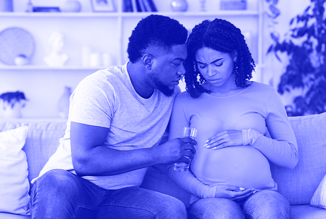 pregnant black woman sits on couch and touches her belly and looks at it in concern next to black man who offers her glass of water