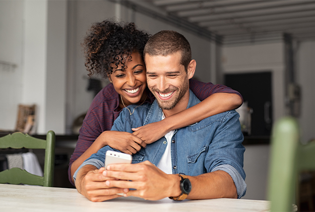 woman smiles draping herself over the shoulders of man sitting and smiling on his phone