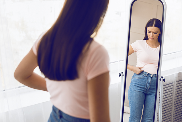 College-age girl in jeans and white tee stands in front of full length mirror and pokes at her side 