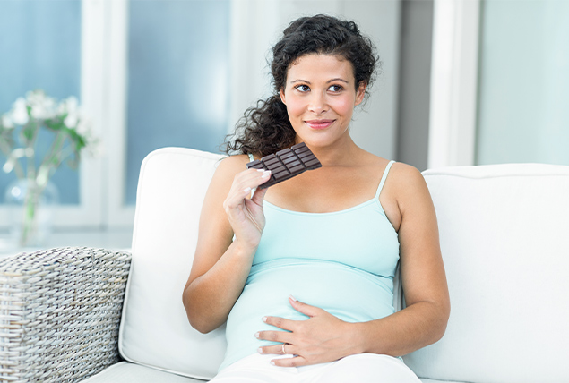 pregnant woman sits on white couch with chocolate bar in hand