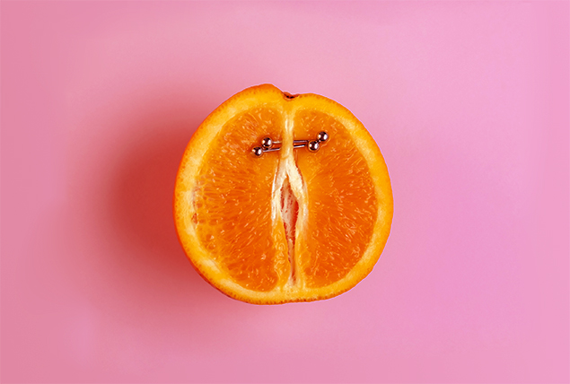 Half of an orange is lying against a pink surface with two barbels piercing through the top center area.