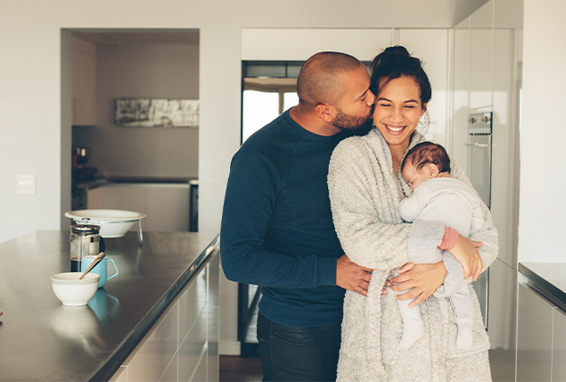 A mother holds their baby while her partner hugs and kisses her cheek.