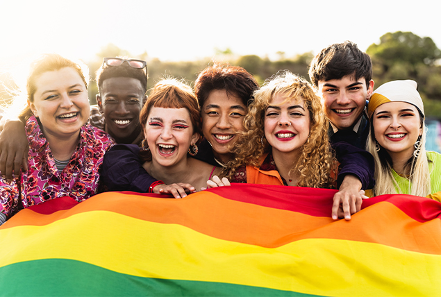 A group of people hold a large rainbow flag together while smiling.