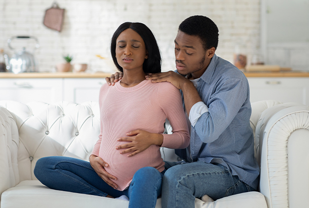 A man sits behind his pregnant partner as she deals with contractions while they sit on the sofa.